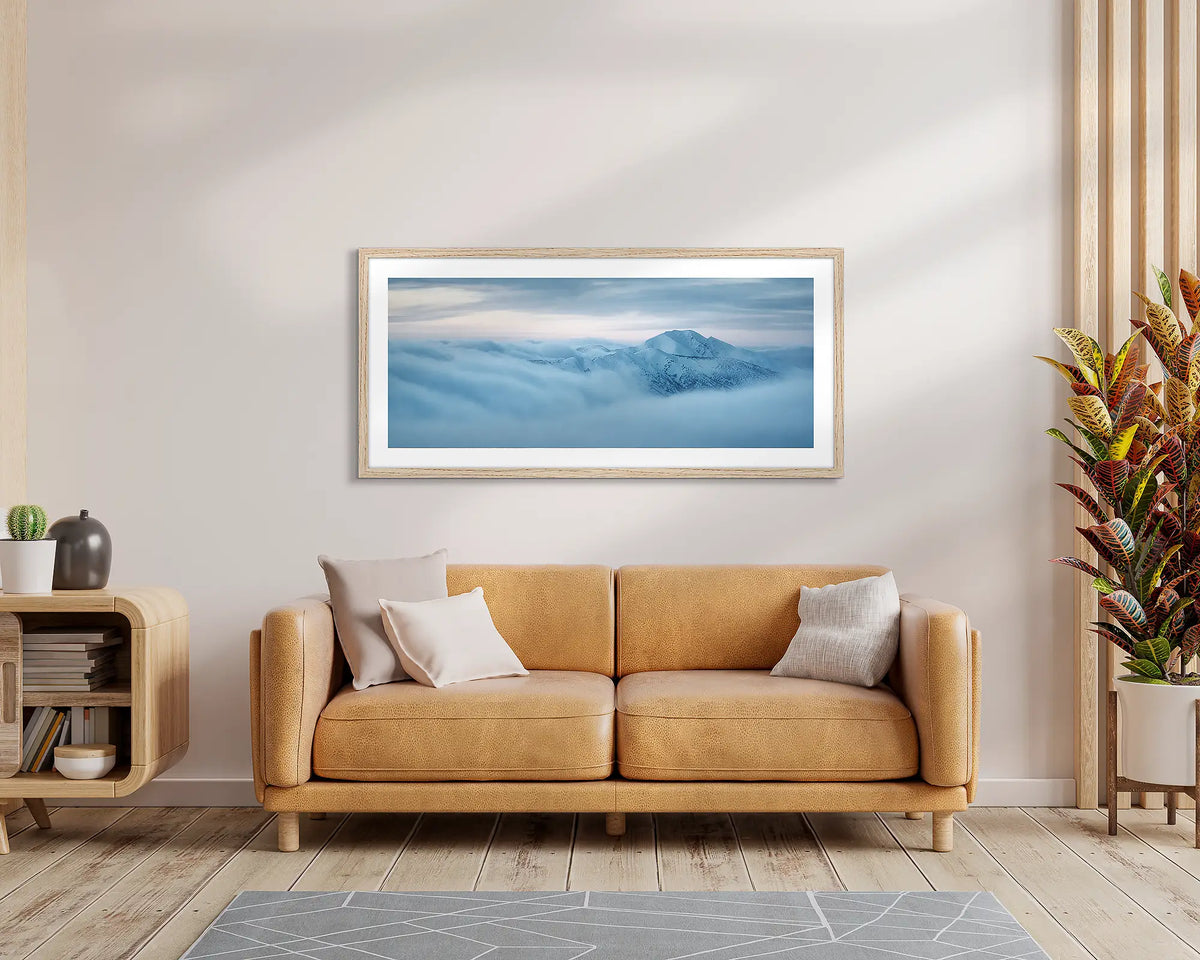 Above It All. Wall art print of Mount Feathertop surrounded by clouds, with a timber frame, hung above a leather couch. 