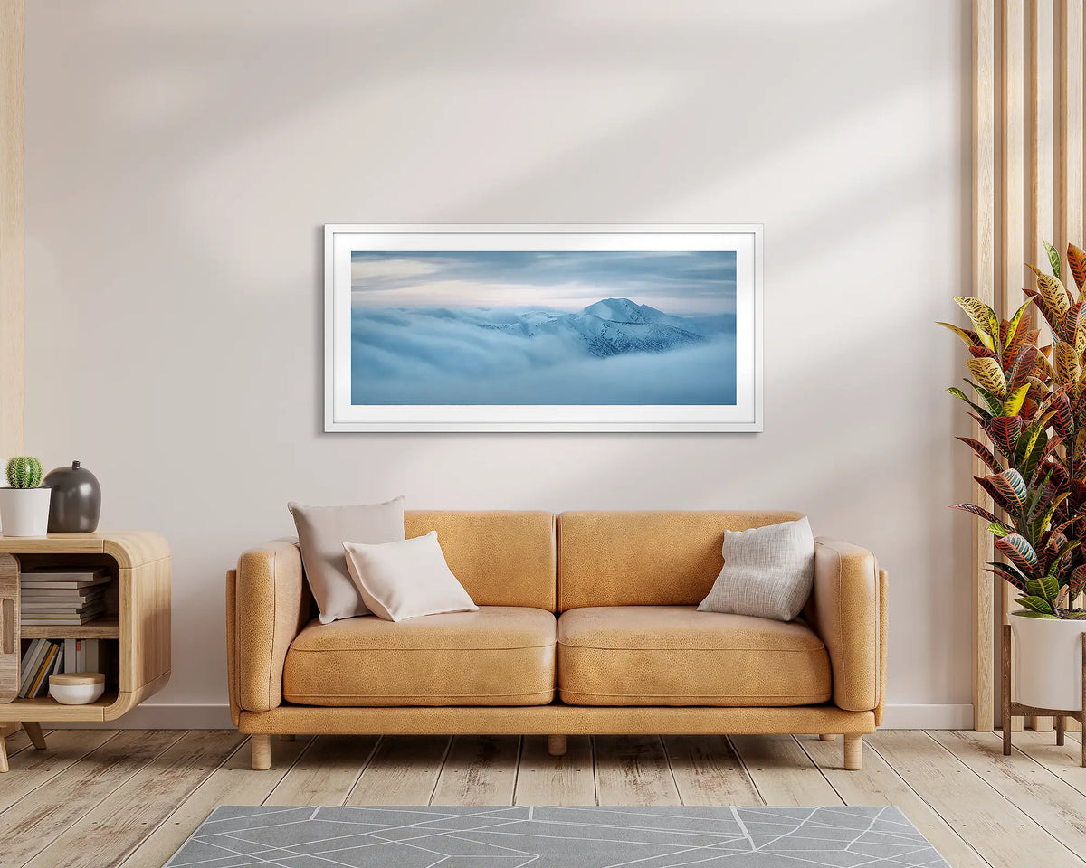 Above It All. Wall art print of Mount Feathertop surrounded by clouds, with a white frame, hung above a leather couch. 