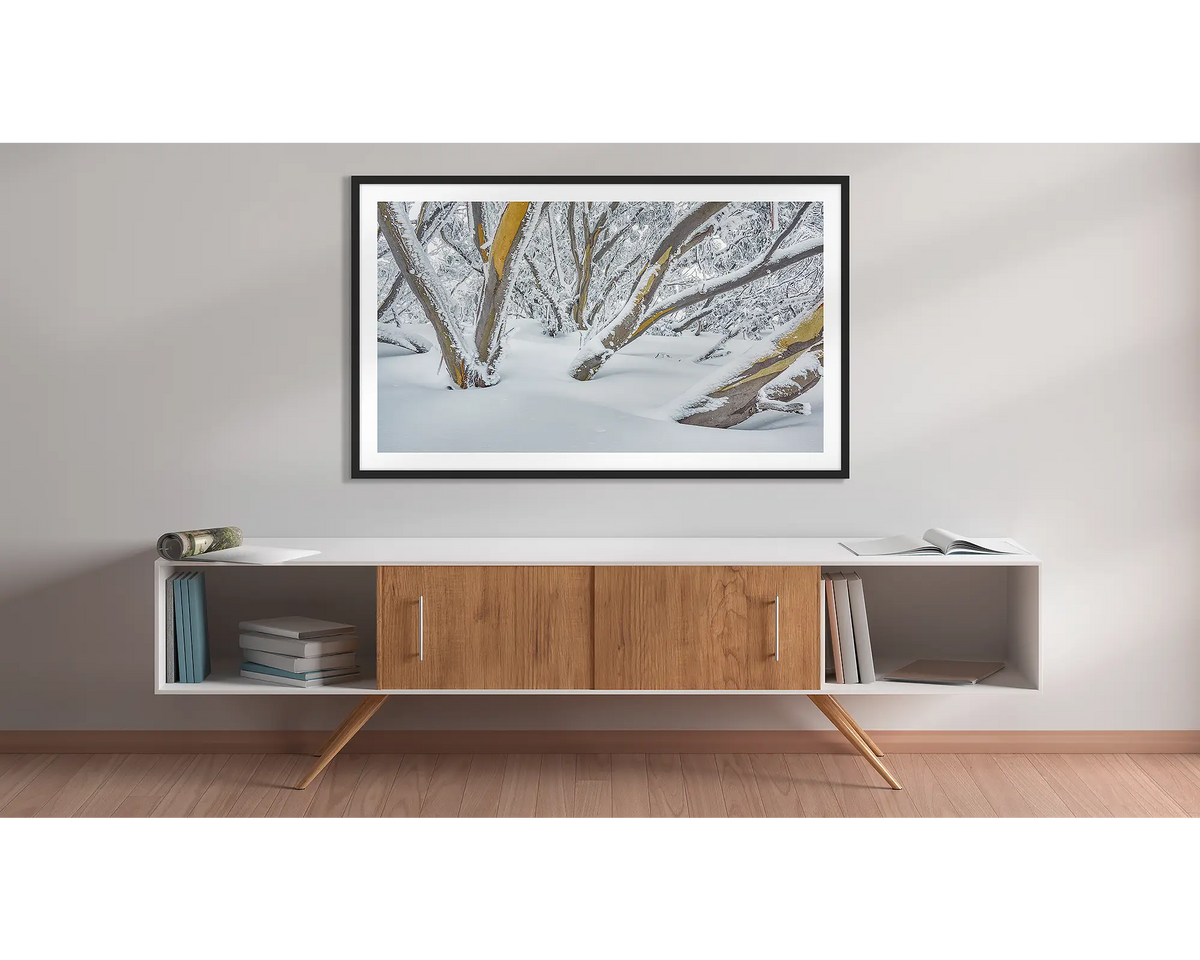 After the Storm. Wall art print of snow gums on Mount Hotham, covered in snow, framed with a black frame, and hung above a side table. 