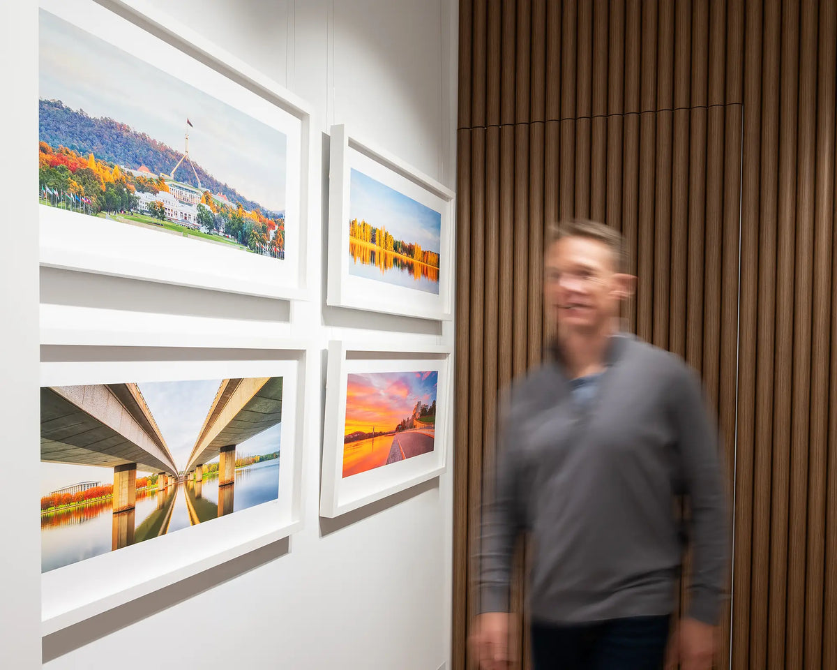 Autumn Bubble. Sunset over Parliament House with white frame, hung in gallery with more Canberra artworks, with person walking beside.