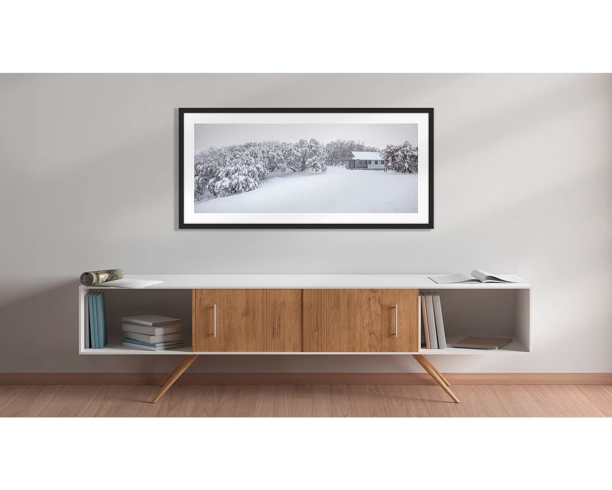 Awaken. Federation Hut with fresh snow wall art print with a black frame hung above a side table. 