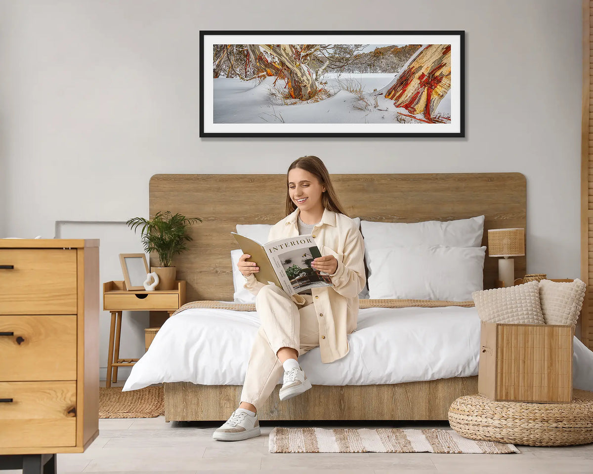 Back Country. Snow gum in snow wall art print, with a black frame, hung above a bed with a woman reading a magazine. 