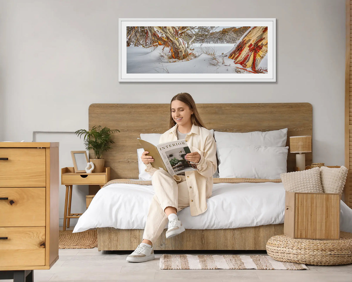 Back Country. Snow gum in snow wall art print, with a white frame, hung above a bed with a woman reading a magazine. 