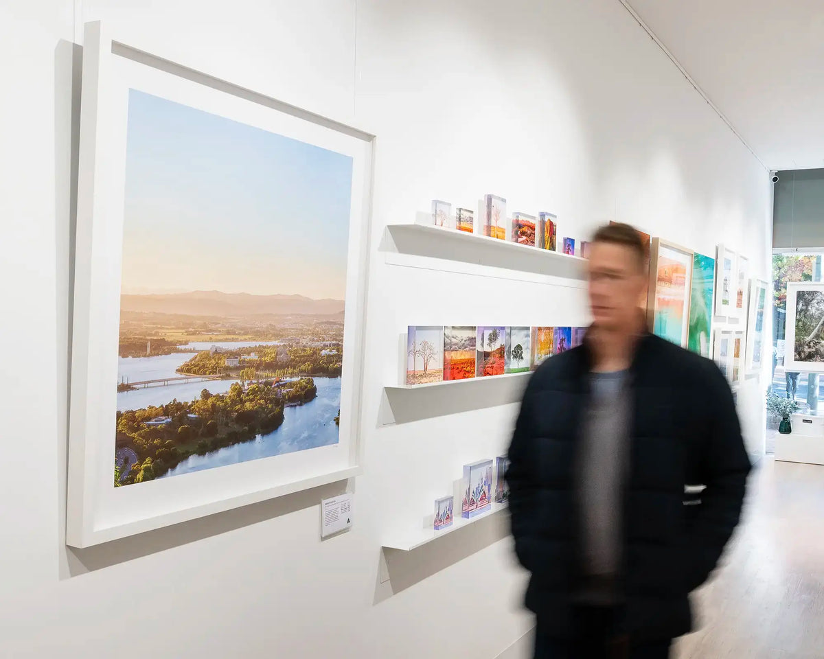 Brighter Days. Bright autumn sunrise over Canberra with white frame, hung in gallery, with person walking beside.