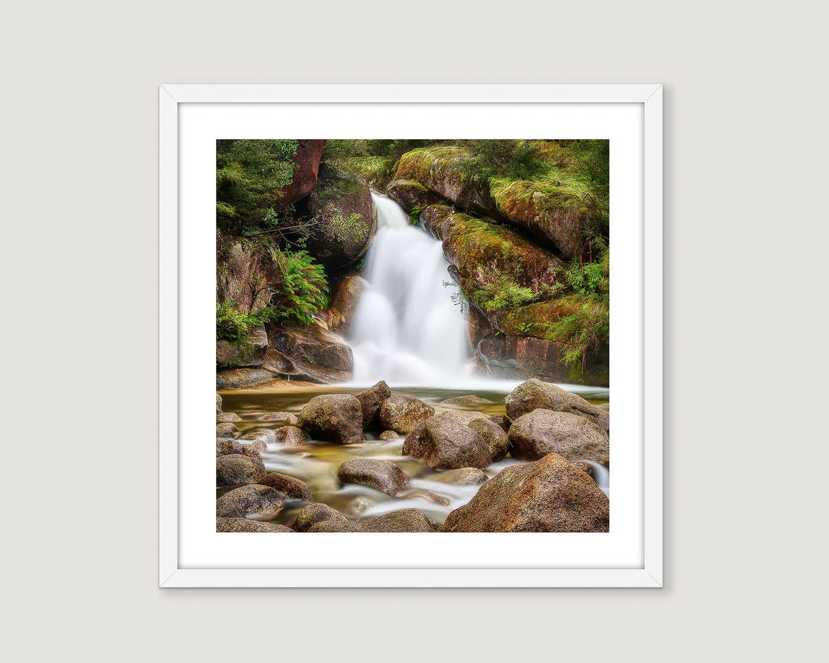 Framed photograph of a waterfall surrounded by rocks and greenery on a white wall.
