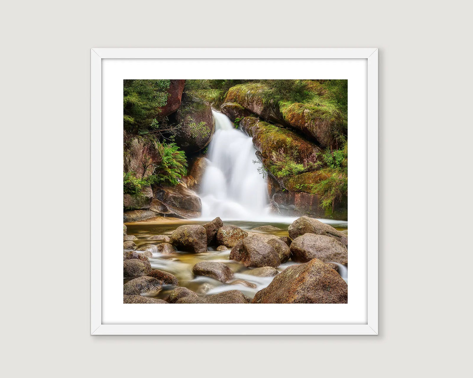 Framed photograph of a waterfall surrounded by rocks and greenery on a white wall.