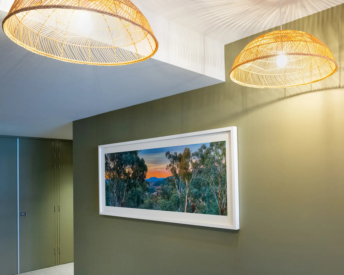 Bush Capital. Canberra view through bushland, with white frame, hung on olive green wall with warm lighting.