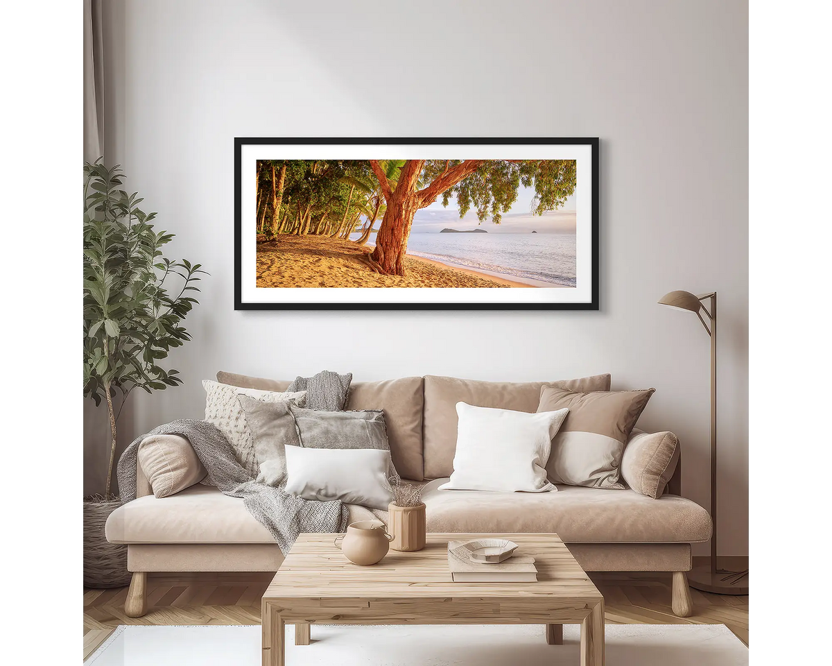 Cairns Escape. Wall art print of a sunrise on a tropical beach, framed with a black frame, hung above a couch.