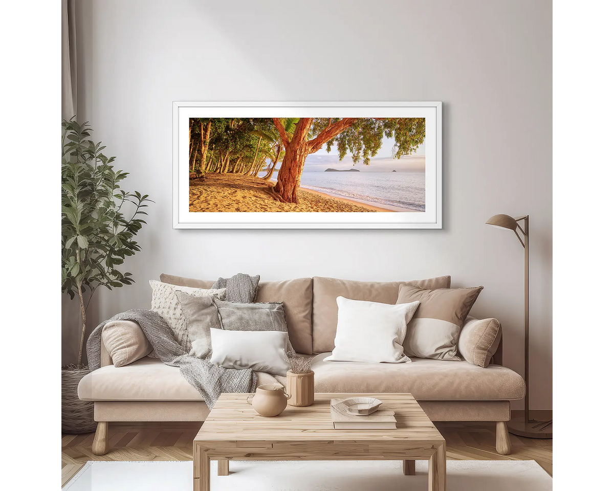 Cairns Escape. Wall art print of a sunrise on a tropical beach, framed with a white frame, hung above a couch.