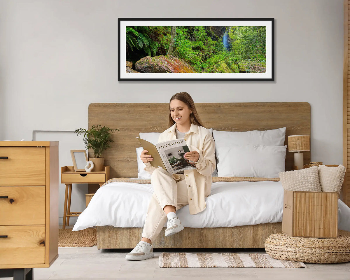 Canyon Escape. Blue Mountains wall art print, with a black frame, hung above a bed with a woman reading a magazine. 