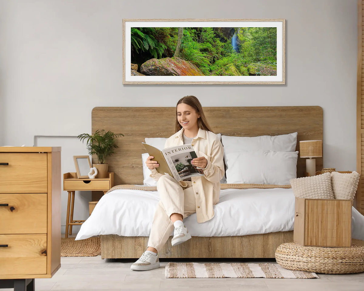 Canyon Escape. Blue Mountains wall art print, with a Tasmanian Oak frame, hung above a bed with a woman reading a magazine. 