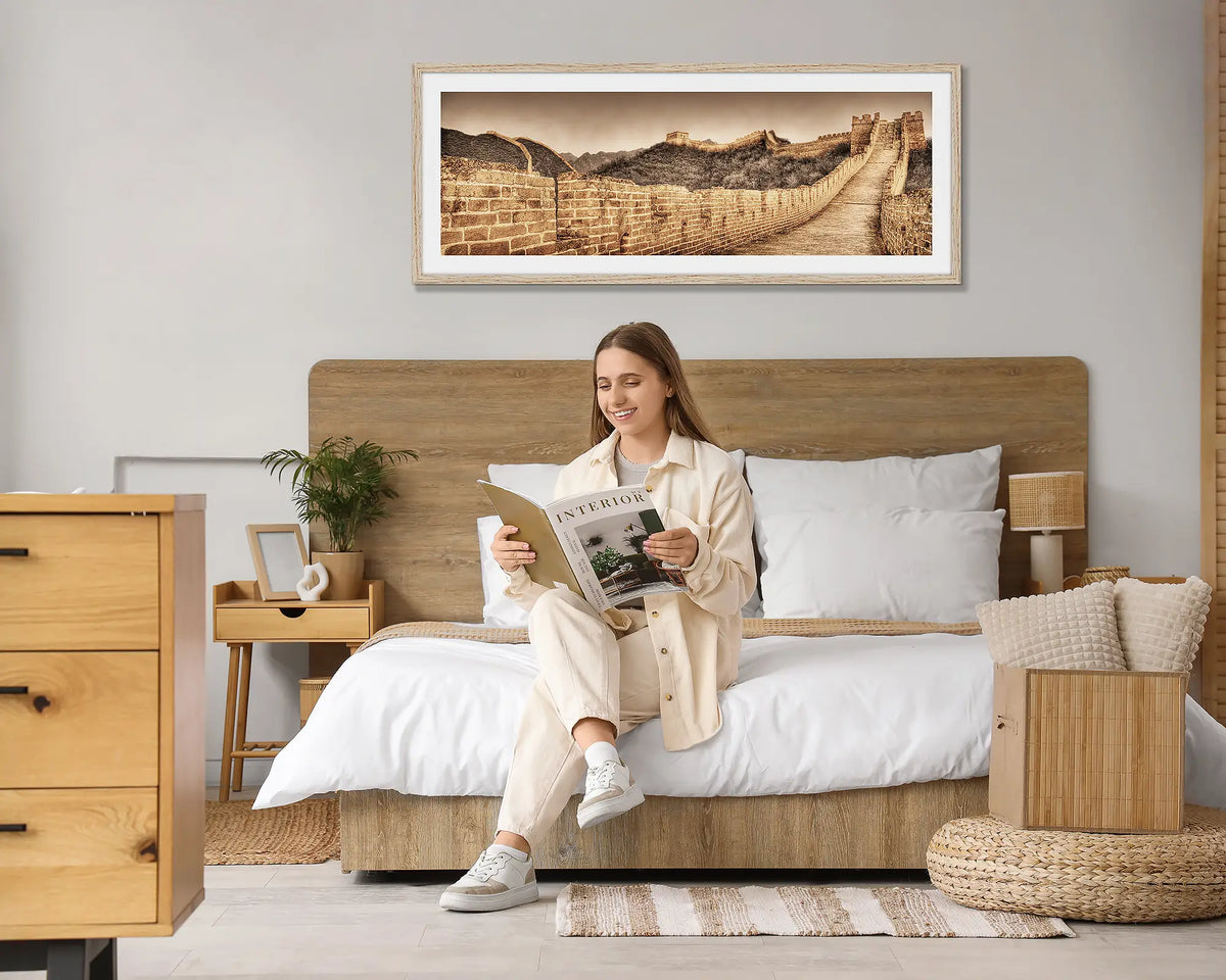Crumbling Wall. Great Wall of China wall art print, with a Tasmanian Oak frame hung above a bed, with a woman reading a magazine. 