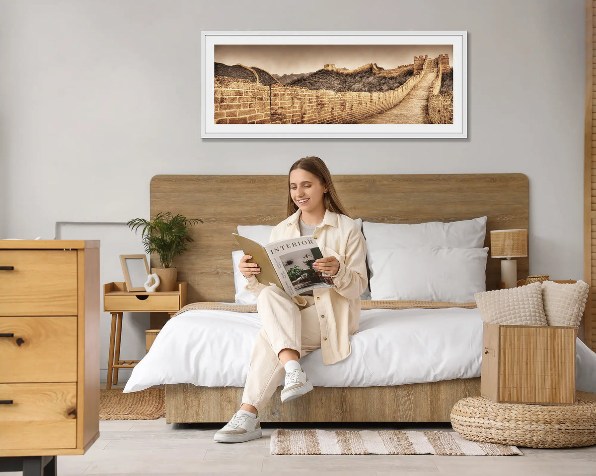 Crumbling Wall. Great Wall of China wall art print, with a white frame hung above a bed, with a woman reading a magazine. 