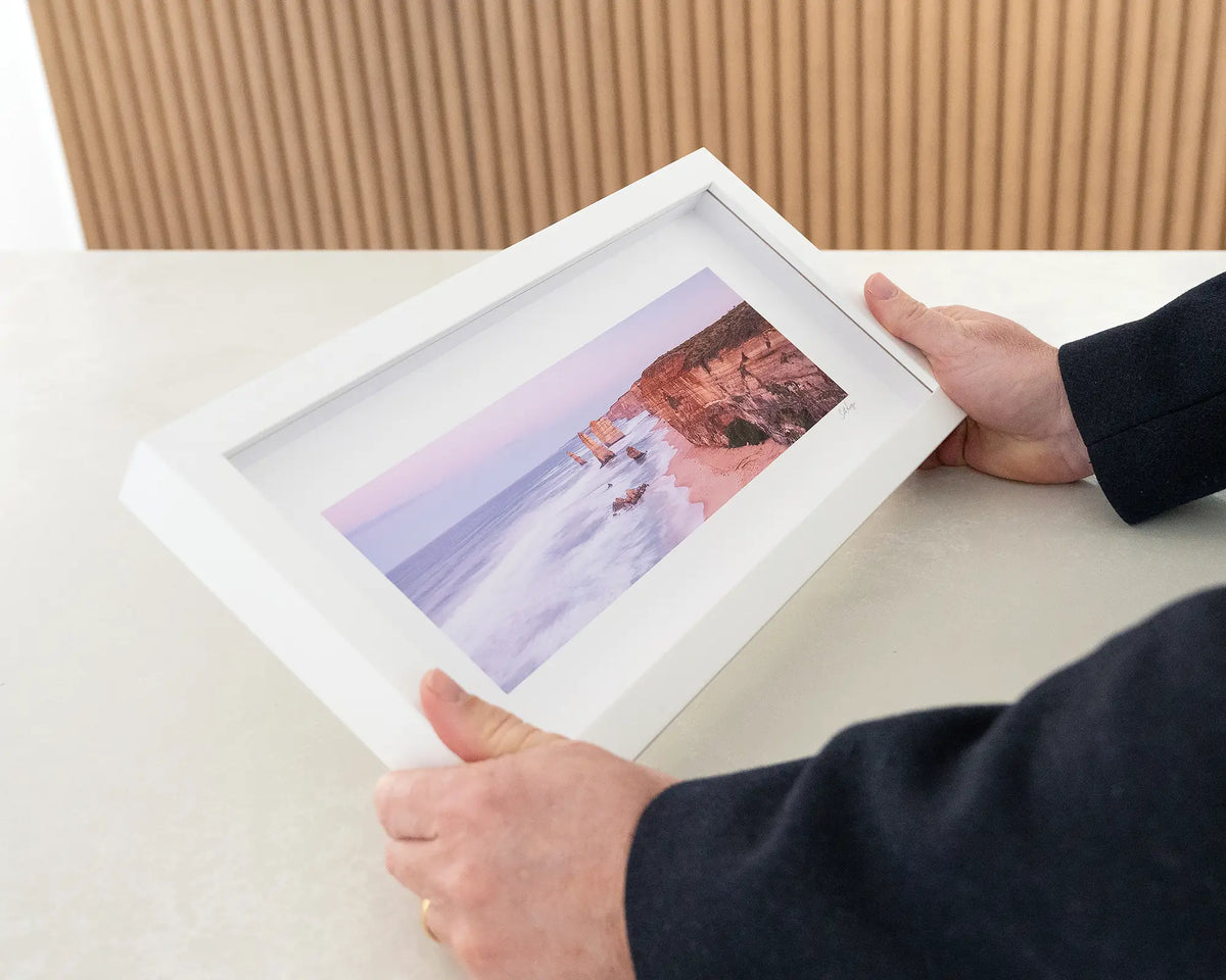 Dawn at the Apostles. Twelve Apostles small framed print with white frame being hand held.