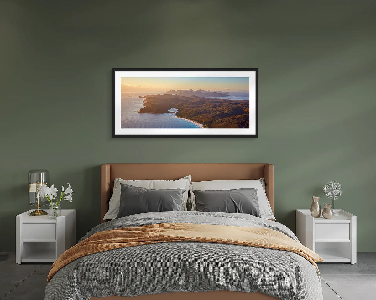 Dawn Over Freycinet. Framed wall art print of a sunrise over Freycinet National Park, with a black frame hung on a green wall above a bed. 