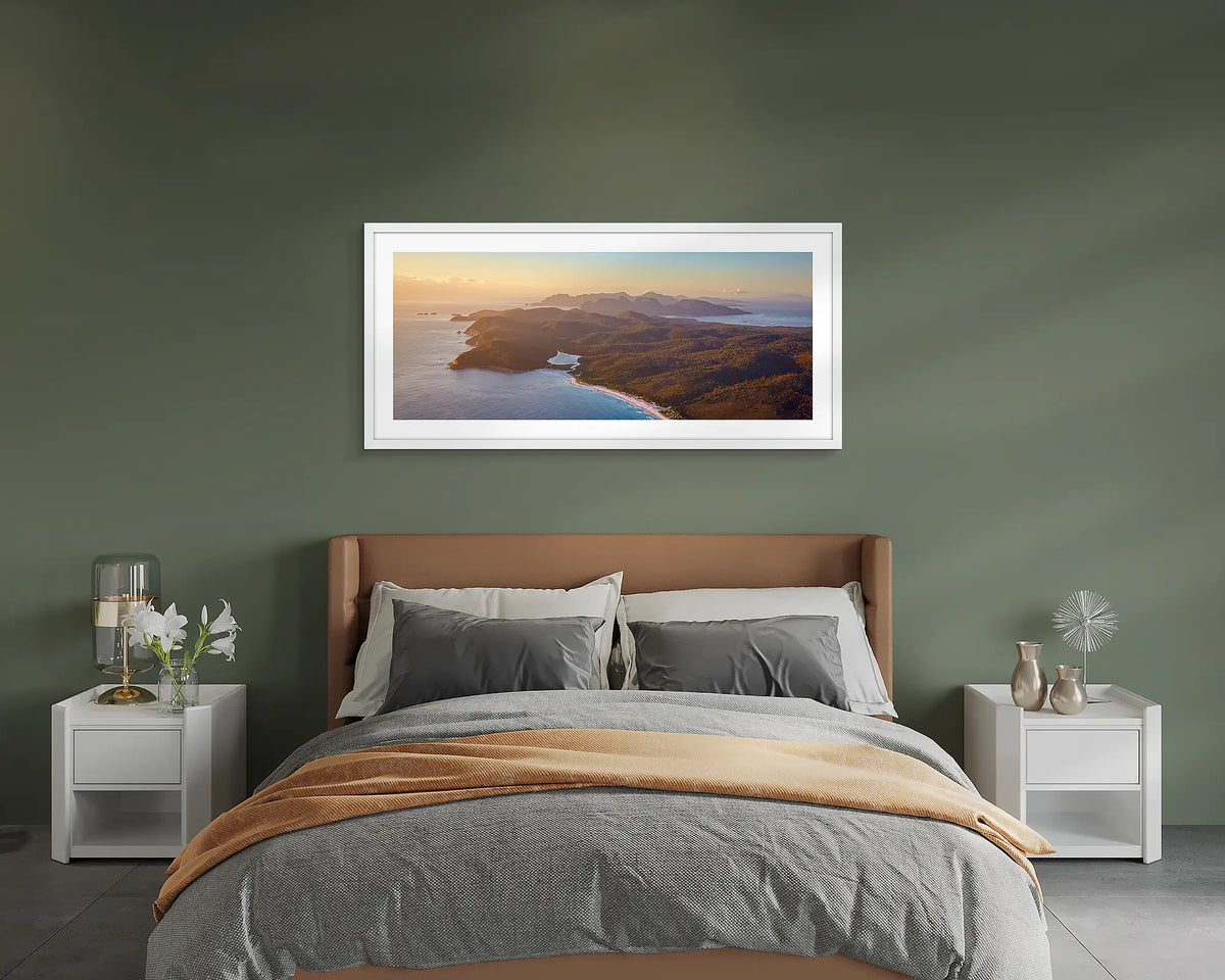 Dawn Over Freycinet. Framed wall art print of a sunrise over Freycinet National Park, with a white frame hung on a green wall above a bed. 