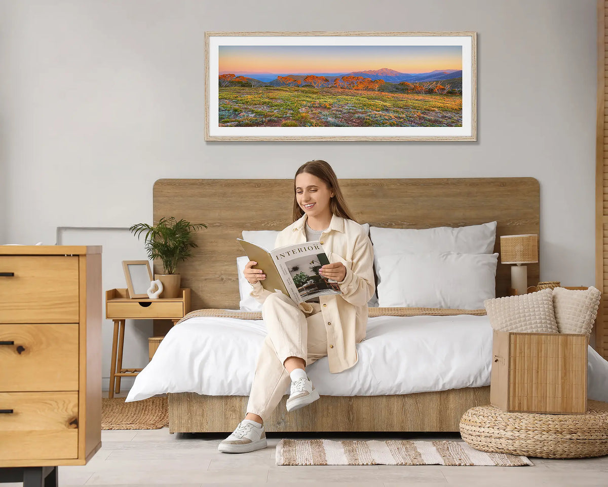 Day Break. Wall art print of a sunrise over the Victorian Alps, with a Tasmanian Oak frame, hung above a bed with a woman reading a magazine. 