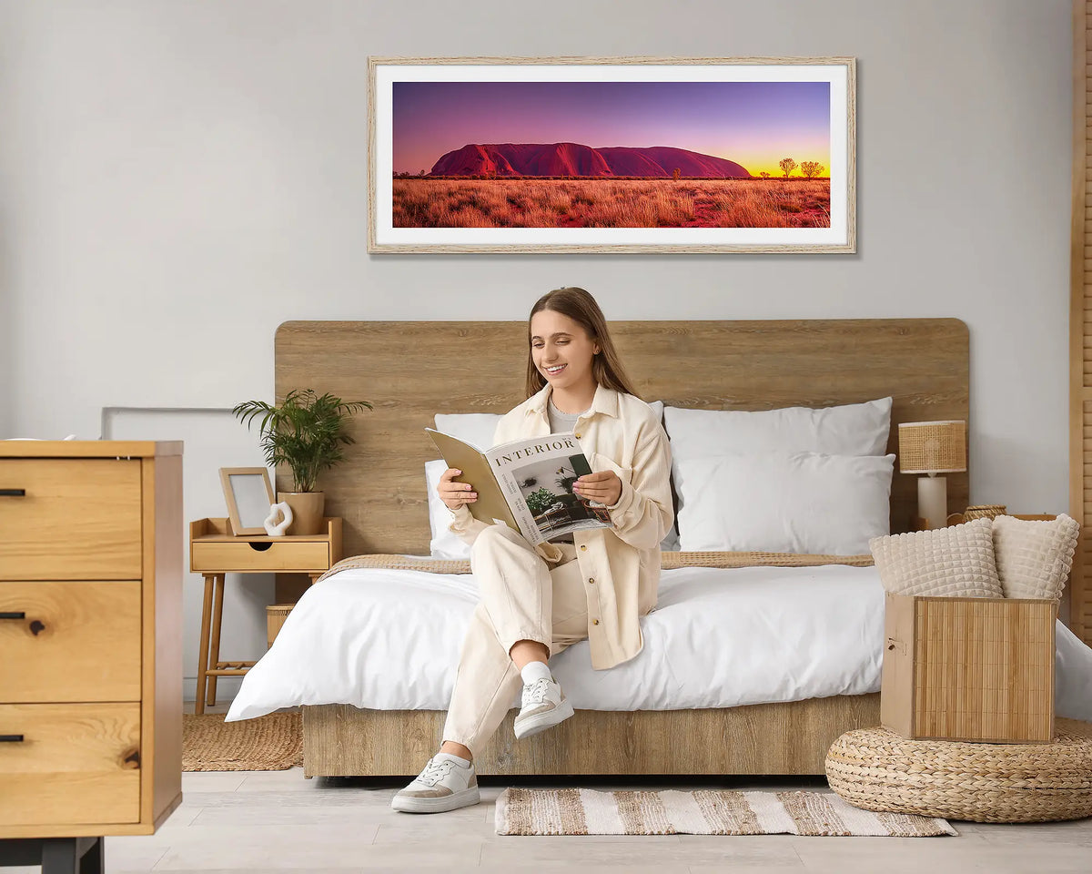 Desert Awakening. Wall art print of a sunrise over Uluru, with a Tasmanian Oak frame, hung above a bed with a woman reading a magazine. 