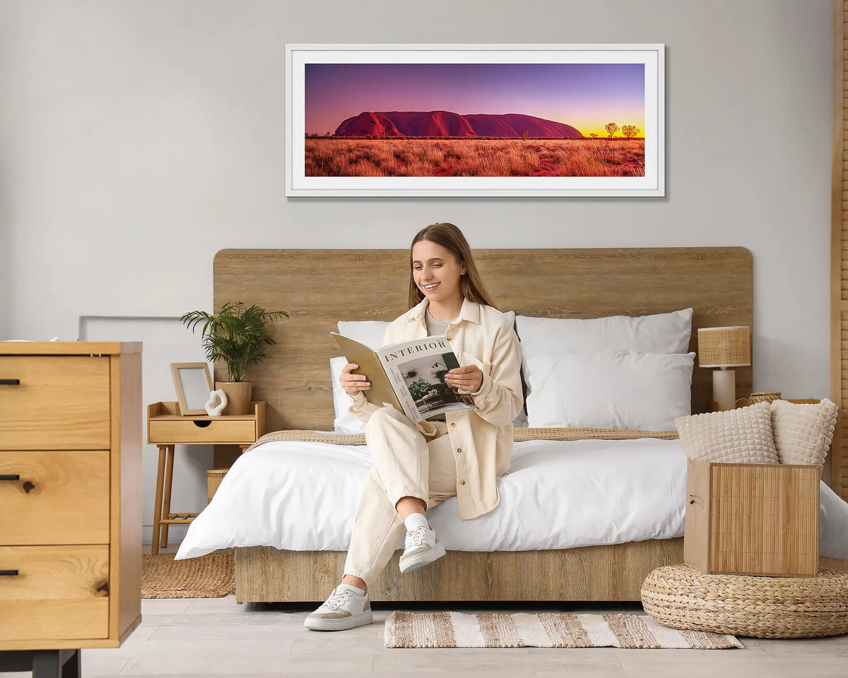 Desert Awakening. Wall art print of a sunrise over Uluru, with a white frame, hung above a bed with a woman reading a magazine. 