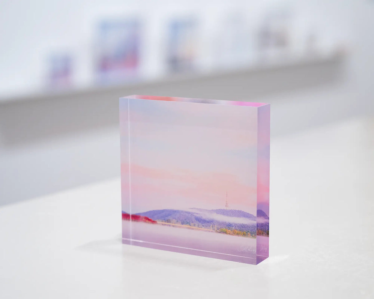 Dreamy Canberra. Acrylic block soft pink sky over Lake Burley Griffin, on gallery desk with display shelf behind.