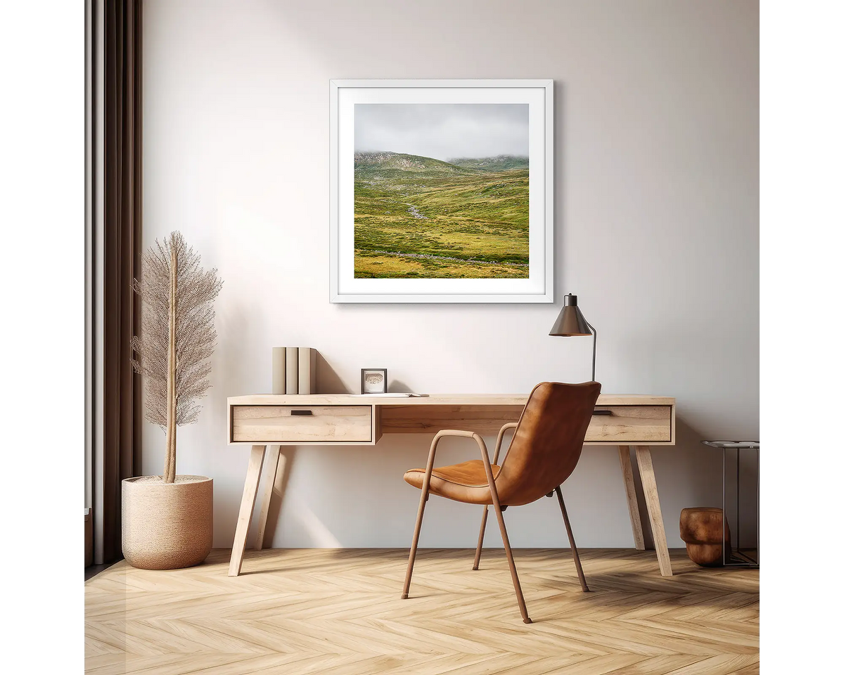 Drizzle. Framed wall art print of Snowy River, Kosciuszko National Park,  with a white frame in an apartment, above a wooden desk. 