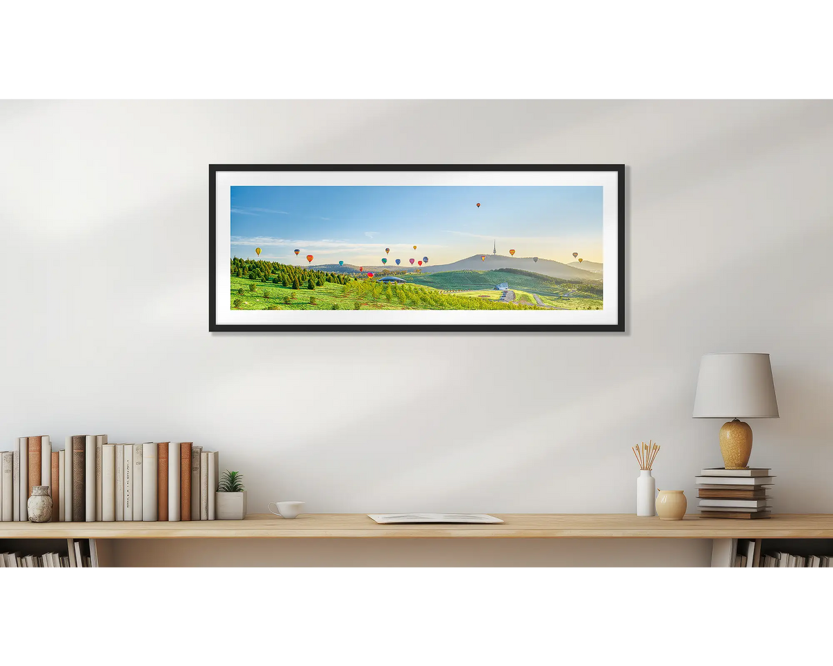 Enlighten. Wall art print of hot air balloons over the arboretum at sunrise, framed with a black frame and hung above a small desk. 