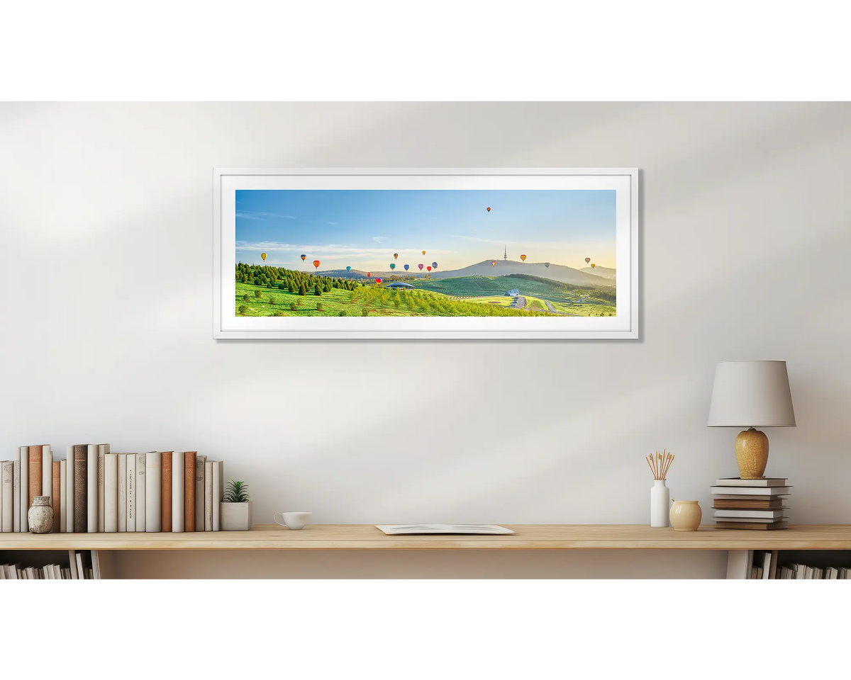Enlighten. Wall art print of hot air balloons over the arboretum at sunrise, framed with a white frame and hung above a small desk. 