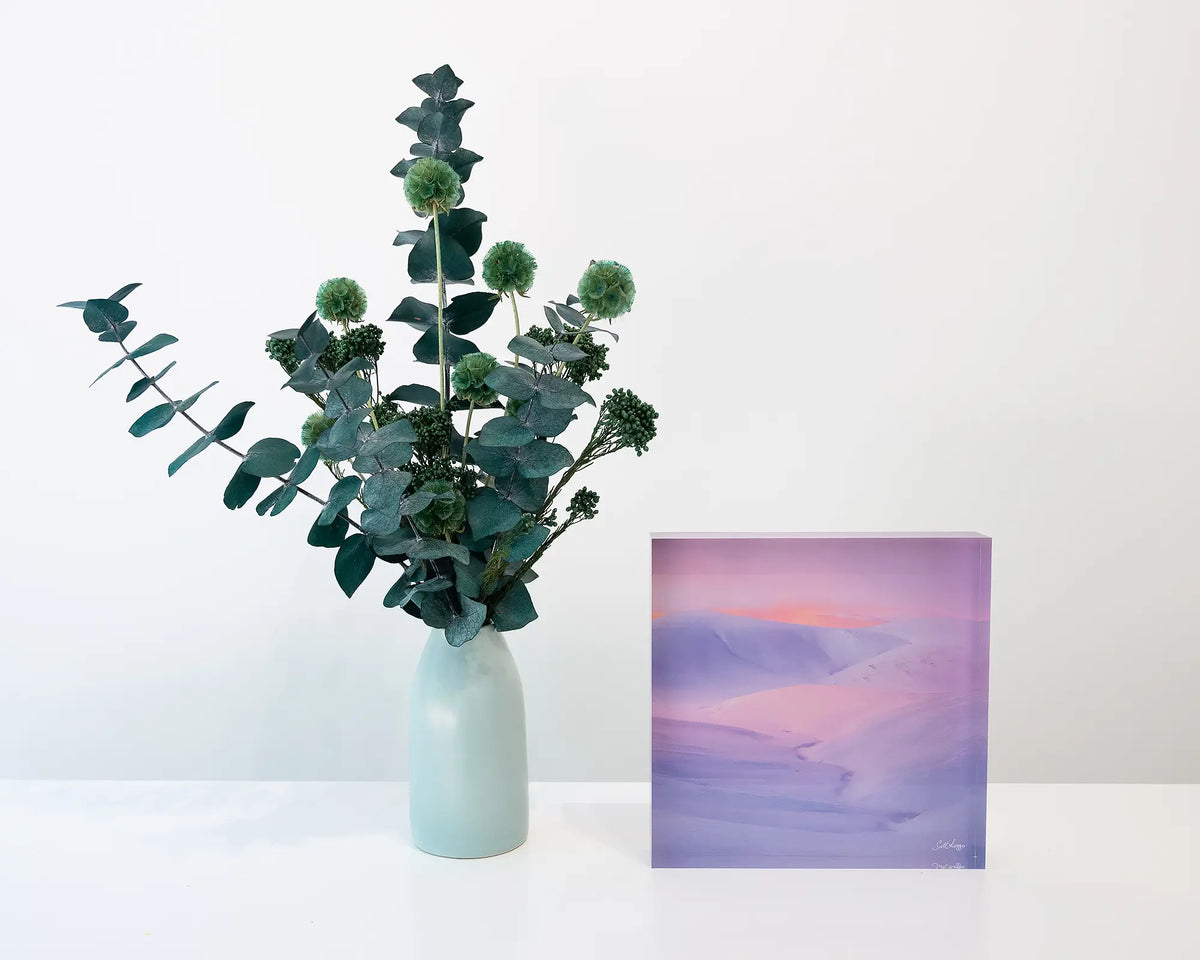Essence Of The Snowy. Acrylic block of sunset over snow covered mountains, Kosciusko National Park, on shelf with floral arrangement.