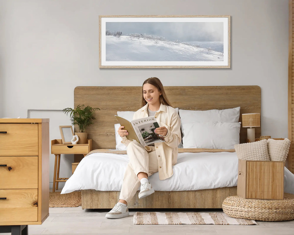 Exposed. Snow wall art print, with a Tasmanian Oak frame hung above a bed, with a woman reading a magazine. 