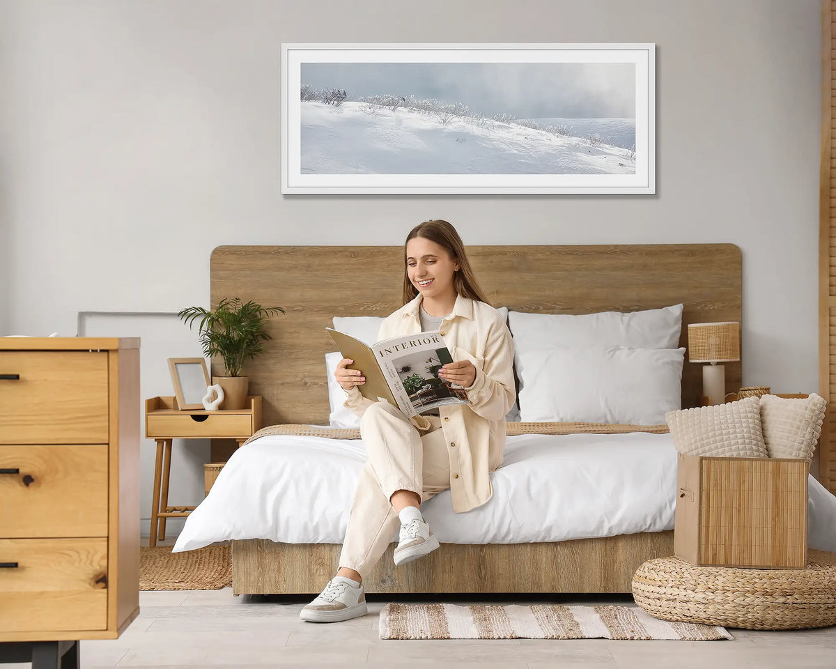 Exposed. Snow wall art print, with a white frame hung above a bed, with a woman reading a magazine. 