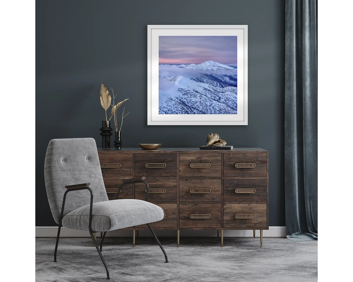 Feathertop Dusk. Wall art print with white frame of Mount Feathertop covered in snow, hung in a room with dark walls.