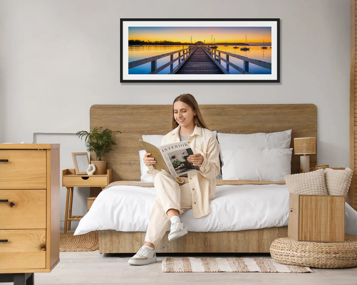 Golden Morning. Batemans Bay sunrise wall art print, with a black frame hung above a bed, with a woman reading a magazine. 