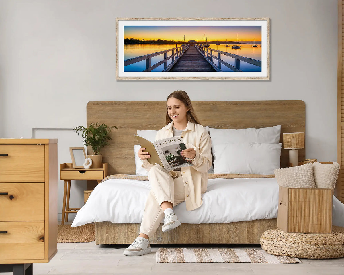 Golden Morning. Batemans Bay sunrise wall art print, with a Tasmanian Oak frame hung above a bed, with a woman reading a magazine. 