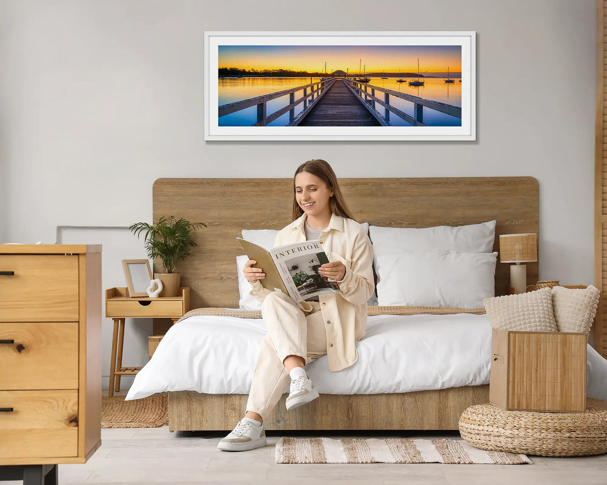 Golden Morning. Batemans Bay sunrise wall art print, with a white frame hung above a bed, with a woman reading a magazine. 