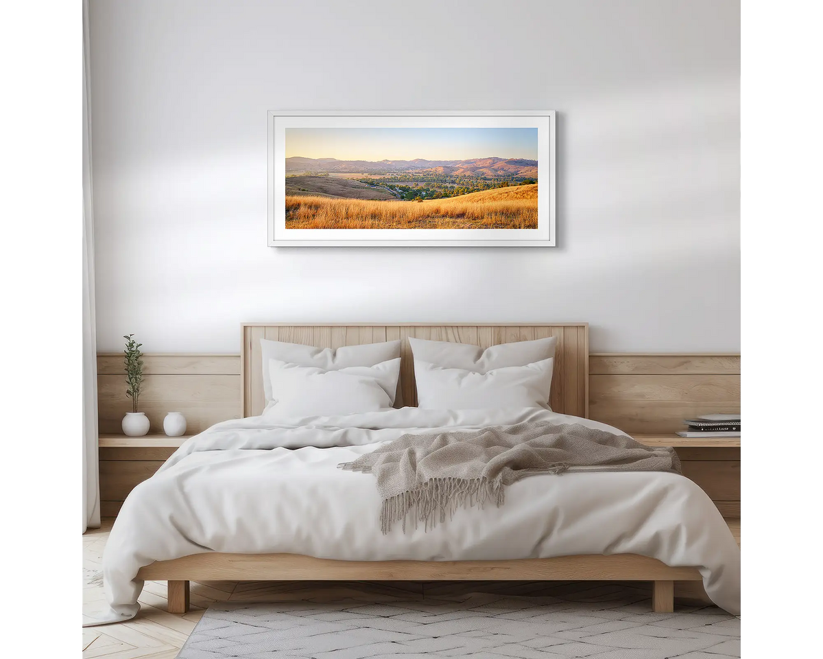 Golden Riverina. Wall art print with a white frame, of a sunrise over Gundagai, hung above a wooden bed. 