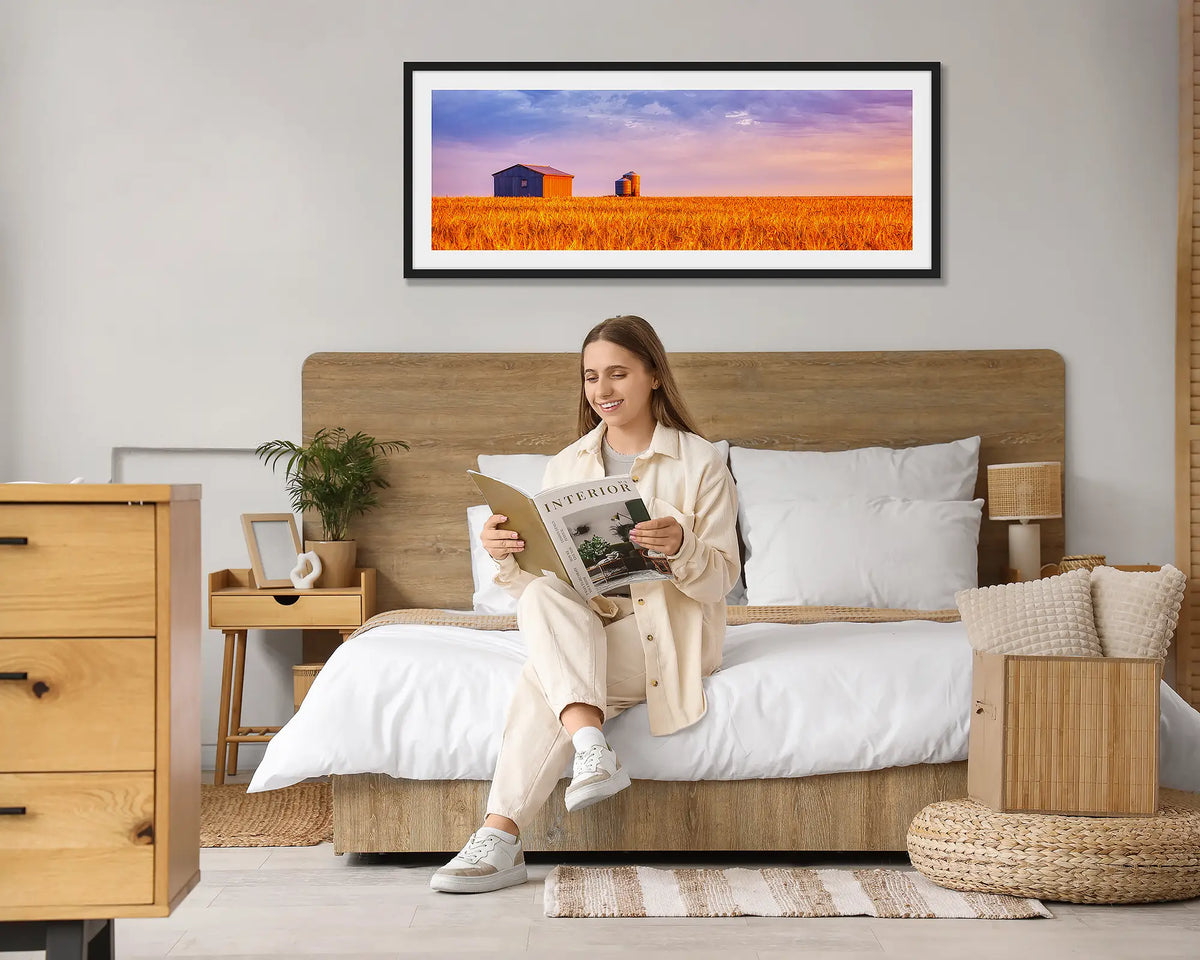 Golden Wheat. Country wheat field wall art print, with a black frame hung above a bed, with a woman reading a magazine. 