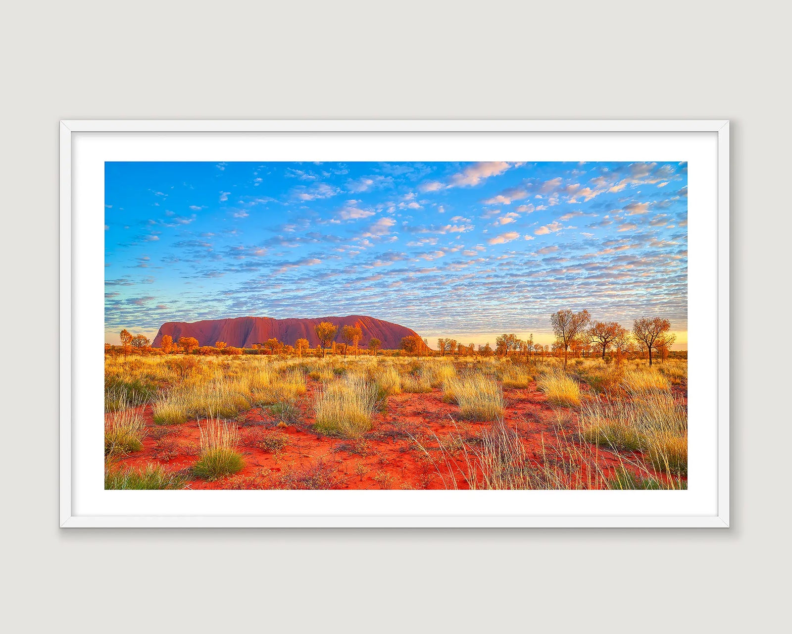 Framed photograph of Uluru with red desert sand, blue sky and scattered clouds.