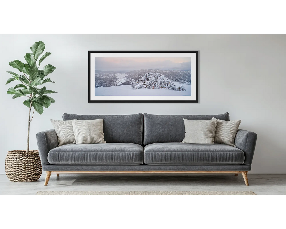 Guardian of the Valley. Wall art print of a snow gum overlooking a snow covered valley, framed in a black frame and hung above a couch. 
