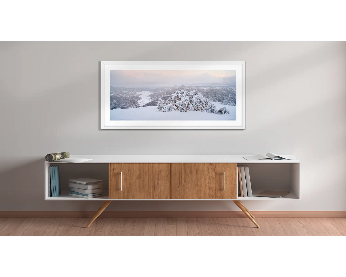 Guardian of the Valley. Wall art print of a snow gum overlooking a snow covered valley, framed in a white frame and hung above a side table. 