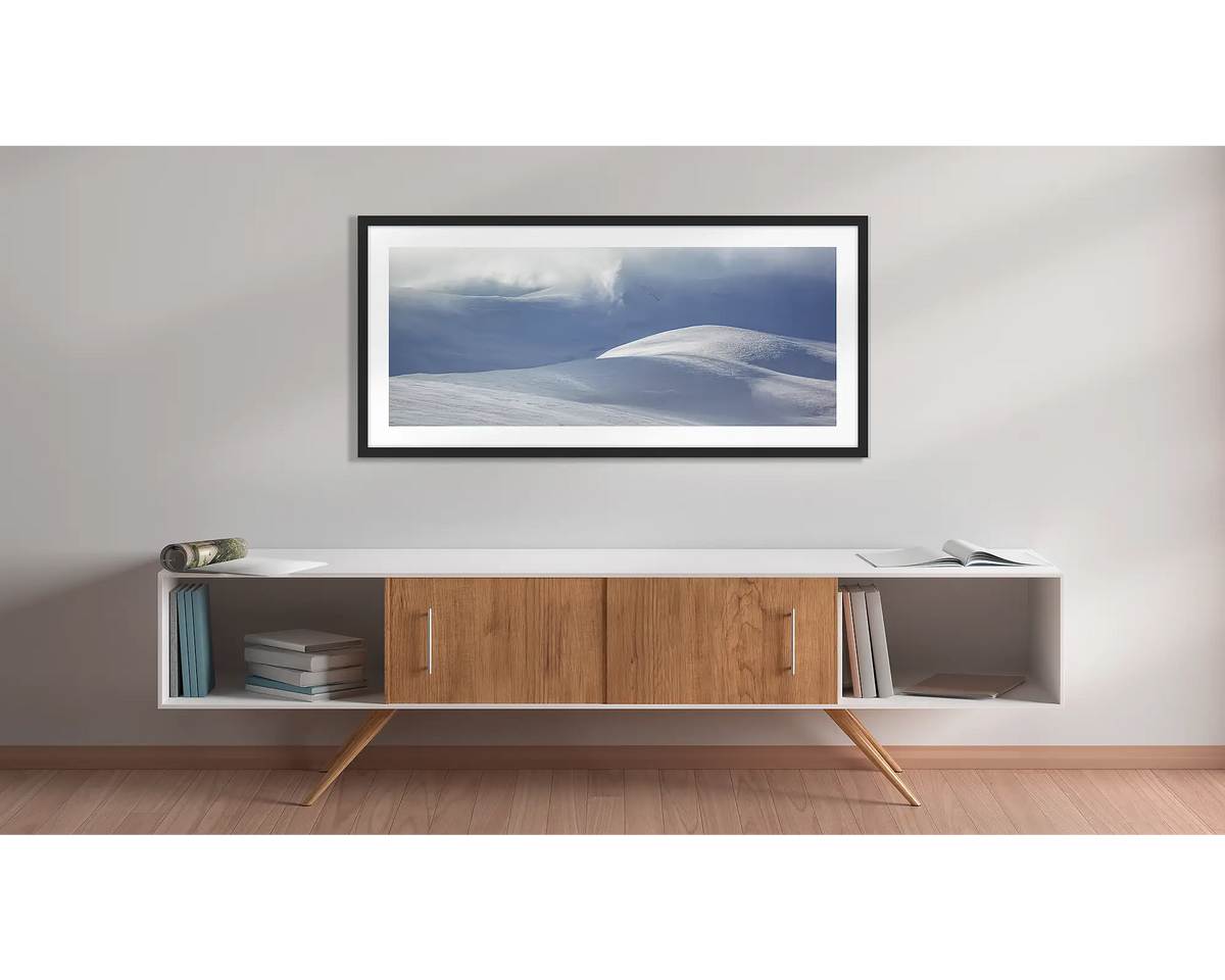 Light Show. Wall art print of light filtering through cloud, onto snow,  framed in a black frame, and hung above a side table. 