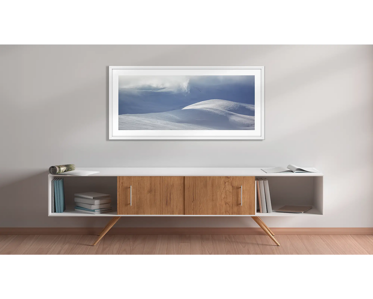 Light Show. Wall art print of light filtering through cloud, onto snow,  framed in a white frame, and hung above a side table. 