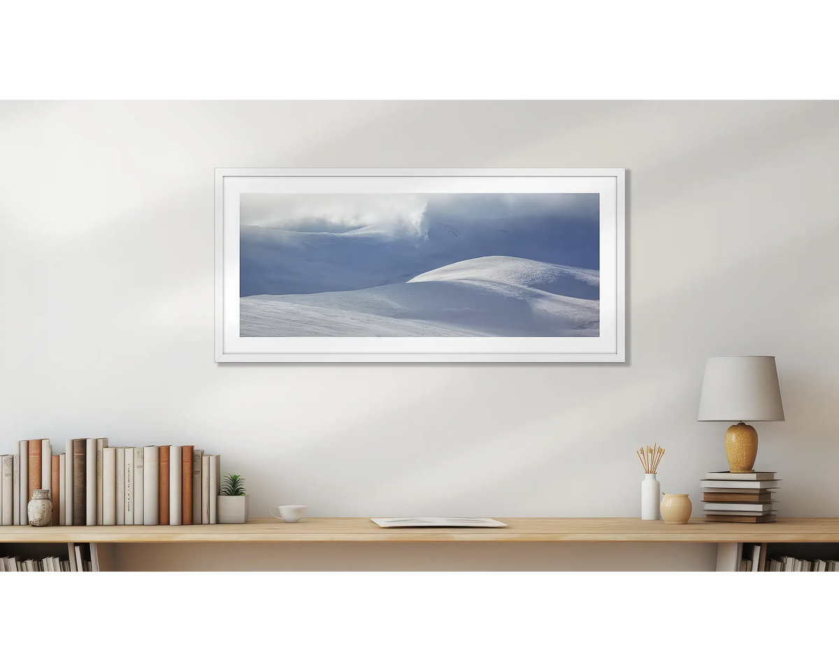 Light Show. Wall art print of light filtering through cloud, onto snow,  framed in a white frame, and hung above a small wooden shelf. 