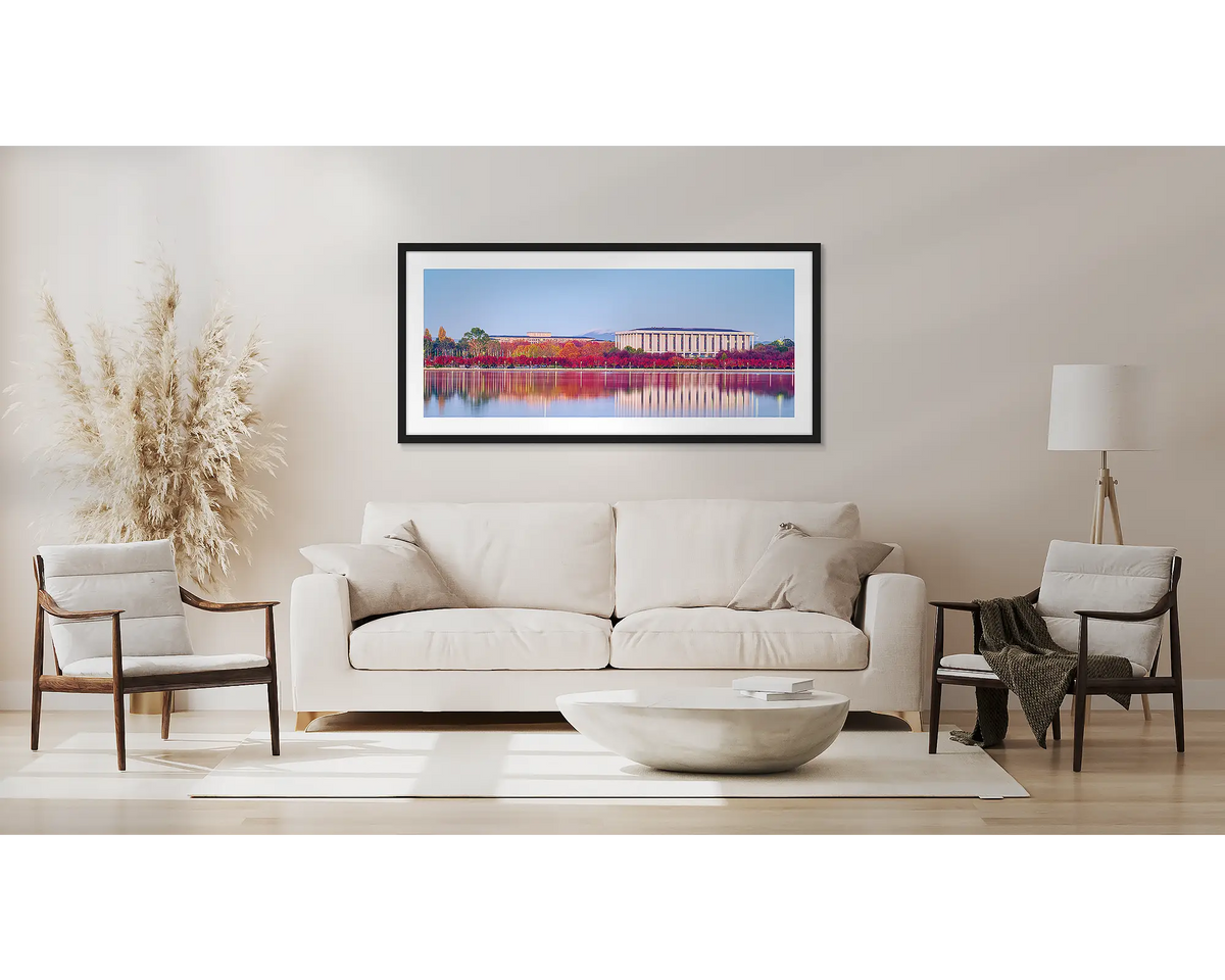 Morning Reds. Wall art print with a black frame hung above a couch, featuring red autumn colours reflecting in the lake, beside the National Library. 