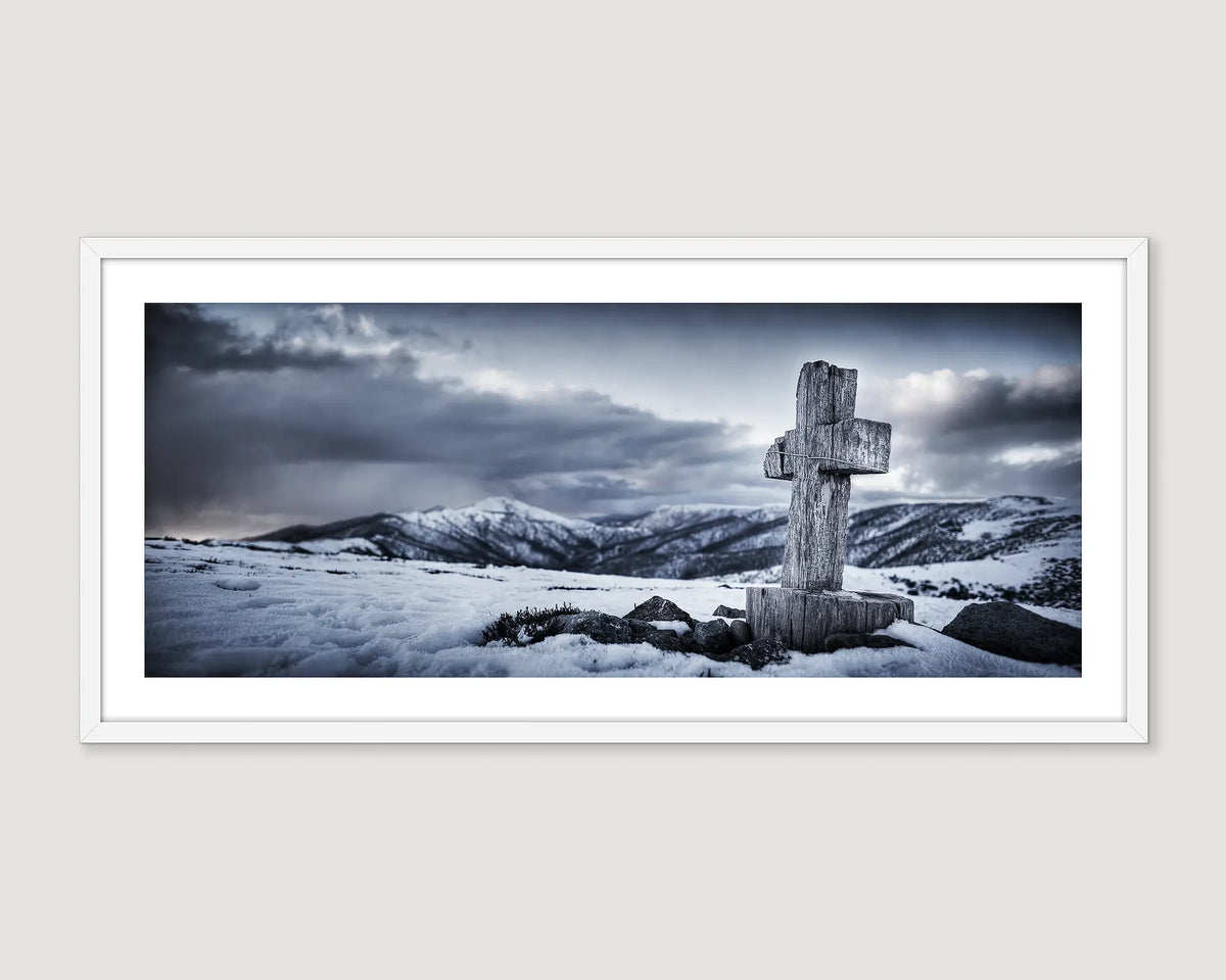 Framed photograph of a cross on a snow covered mountain on a cloudy day.