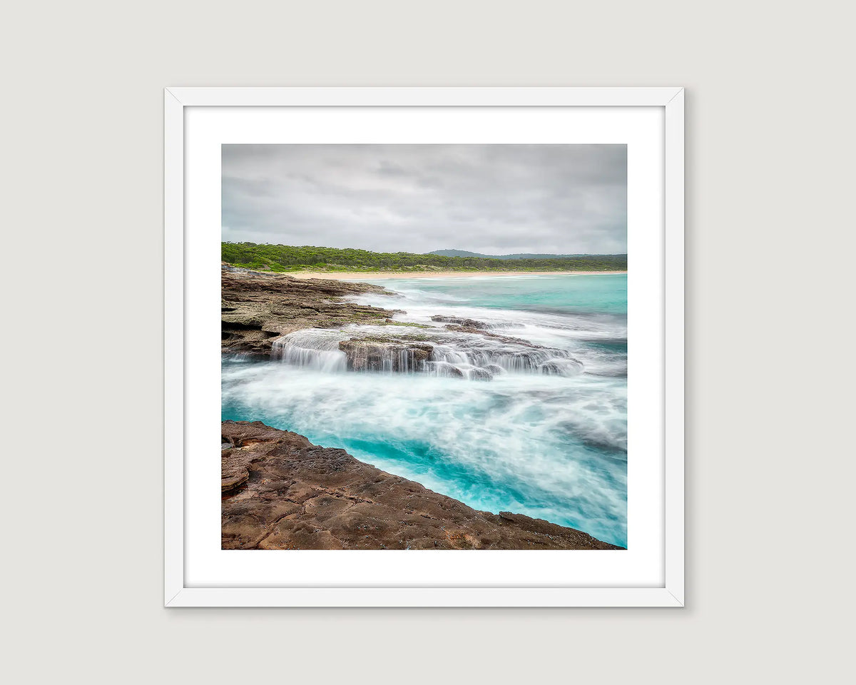 Framed photograph of an ocean flowing over rocks and greenery in the background.