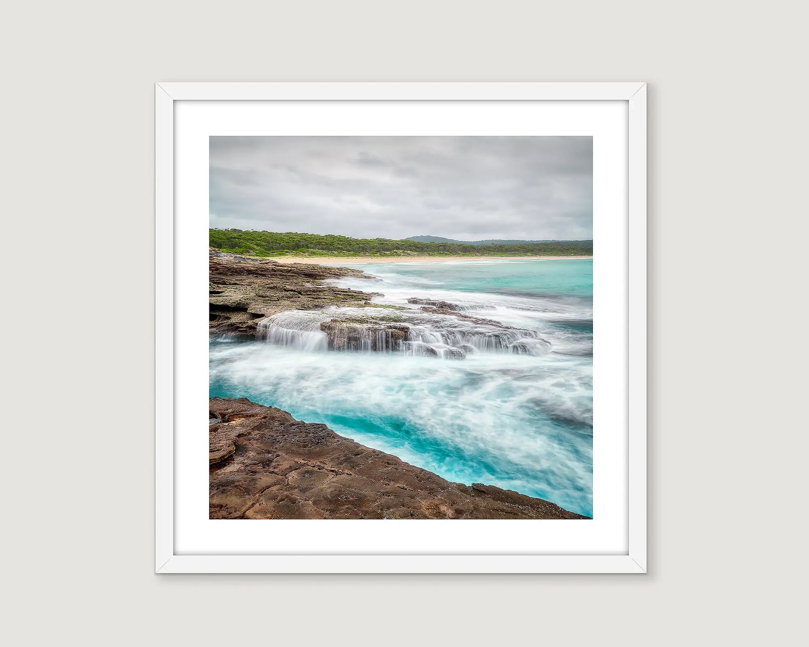 Framed photograph of an ocean flowing over rocks and greenery in the background.