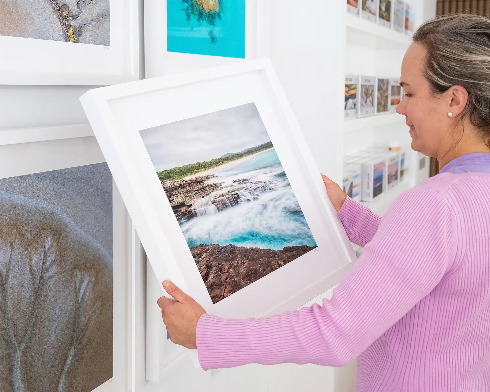 Framed photograph of an ocean flowing over rocks and greenery in the background.