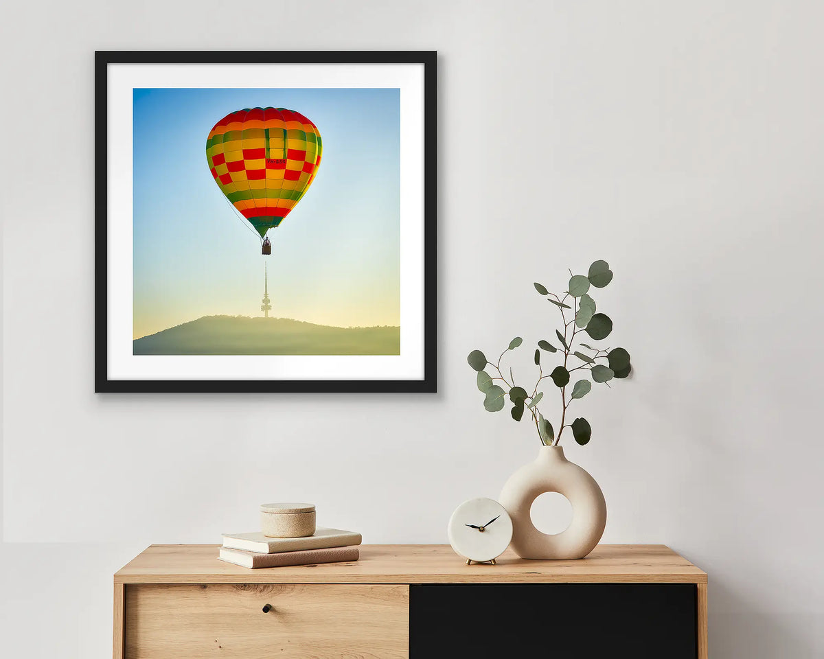 On Top. Canberra wall art print of a hot air balloon and Black Mountain Tower, framed in a black frame and hung above a side table. 