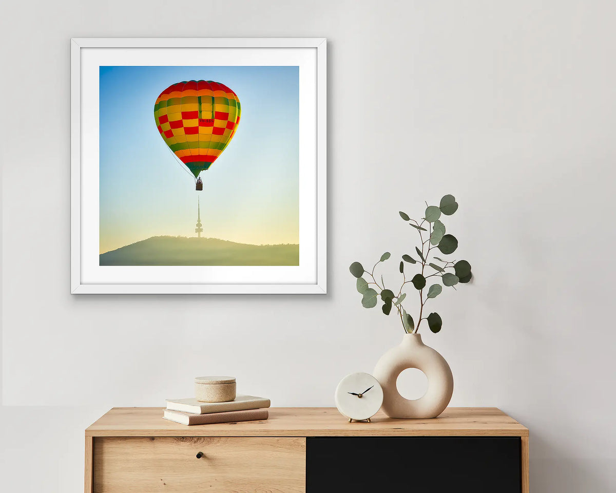 On Top. Canberra wall art print of a hot air balloon and Black Mountain Tower, framed in a white frame and hung above a side table. 
