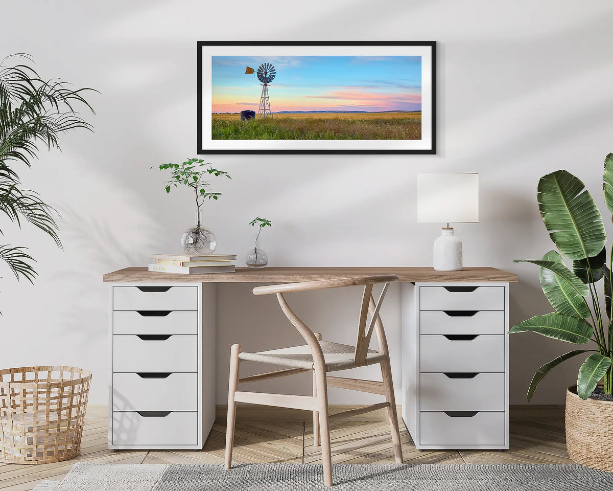Painted Morning. Wall art print of a windmill on a farm, with a colourful sunrise, with a black frame hung above a small desk. 